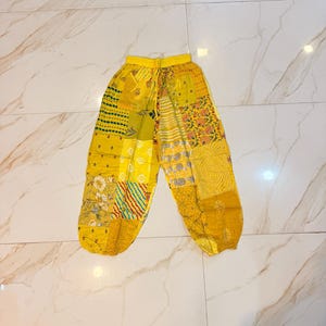 May include: Yellow patchwork pants featuring a variety of printed patterns including floral, striped, and geometric designs. The trousers have an elasticized waistband with a drawstring and elastic cuffs at the ankles, constructed from multiple fabric panels.
