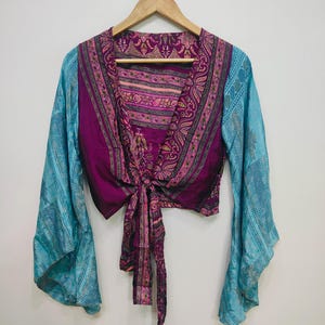 May include: A long-sleeved wrap top featuring a deep purple bodice with a vibrant pink and gold floral pattern. The garment includes wide, flowing sleeves in a contrasting light blue fabric with a subtle, intricate geometric print throughout.