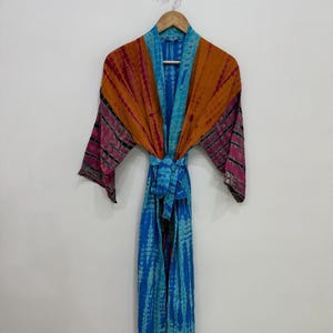 Könnte beinhalten: Ein langärmeliger Morgenmantel mit einem Batikmuster in Blau-, Orange- und Magentatönen. Das Kleidungsstück verfügt über einen passenden Stoffgürtel in der Taille, einen breiten Kragen und eine offene Vorderseite, präsentiert auf einem Holzkleiderbügel.