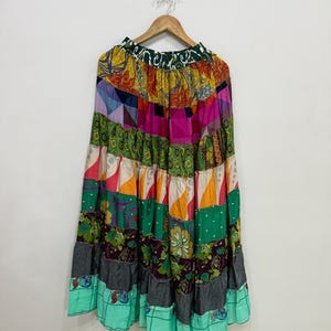 Puede incluir: Una falda patchwork colorida con un diseño escalonado, colgada de una percha de madera. La falda muestra una variedad de telas en tonos verdes, rosas, naranjas y turquesas. El diseño de la falda es bohemio y único, con una mezcla de patrones y texturas.