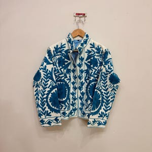 以下が含まれることがあります： クリーム色のジャケットに、鮮やかなティールブルーの花柄刺繍が施されています。 ジャケットはハイカラーで、木製のハンガーに掛けられています。 刺繍はクリーム色の生地とのコントラストが際立ち、詳細な植物のデザインを示しています。