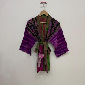 May include: A colorful, patterned robe with a tie belt. The robe features a mix of purple, green, and black hues. The sleeves are wide and the fabric appears to be lightweight. The robe is hanging on a wooden hanger against a white wall.