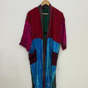 Puede incluir: Una bata tipo kimono de manga larga con un diseño de bloques de color en rojo oscuro, magenta y azul brillante. La prenda incluye un patrón tie-dye en las áreas azules y magenta, un borde verde oscuro en la abertura frontal y dos bolsillos.