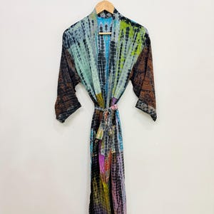 May include: A long, tie-dye robe with a colorful pattern of blues, greens, purples, and browns. The robe is hanging on a wooden hanger against a white wall. The robe has long sleeves and a matching tie belt.