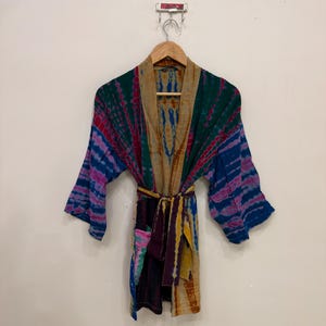 May include: A long-sleeved kimono robe featuring a vibrant tie-dye pattern in shades of deep green, magenta, royal blue, and yellow. The garment includes a matching fabric tie belt at the waist and a tan-colored collar, hanging on a wooden hanger.
