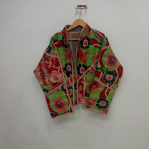 Op de afbeelding: Een doorgestikt jack met een levendig bloemenpatroon in tinten rood, groen en zwart. Het kledingstuk heeft lange mouwen, een kraag en zichtbare witte stiksels langs de randen en naden, hangend aan een houten kledinghanger tegen een muur.
