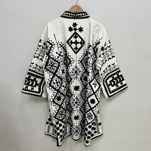 May include: A white cotton tunic with a black geometric embroidery pattern. The tunic has a collar and three-quarter sleeves.