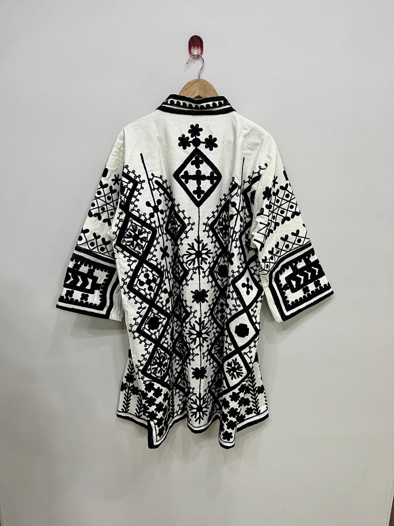 May include: A white cotton tunic with a black geometric embroidery pattern. The tunic has a collar and three-quarter sleeves.