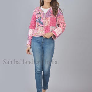 Op de afbeelding: Een roze en witte patchwork gewatteerde jas met bloemenpatronen. De jas heeft een V-hals en lange mouwen met een klokvorm aan de manchetten. Het model draagt ​​een blauwe jeans en staat op een grijze achtergrond.