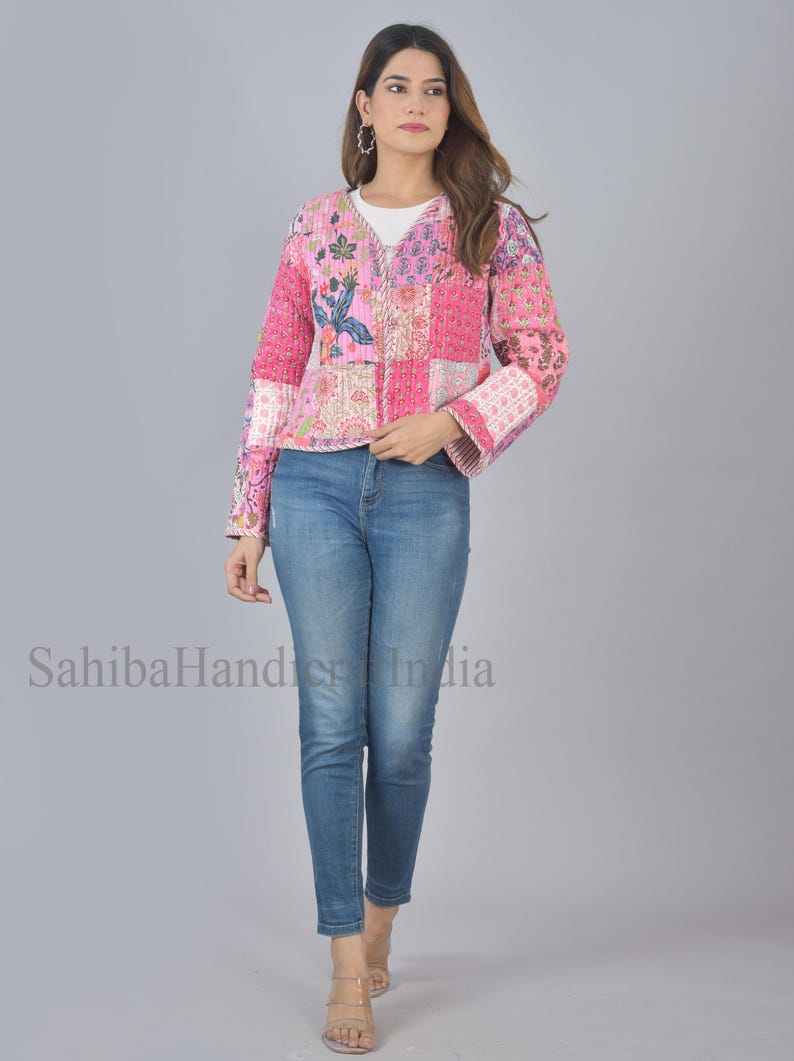 Op de afbeelding: Een roze en witte patchwork gewatteerde jas met bloemenpatronen. De jas heeft een V-hals en lange mouwen met een klokvorm aan de manchetten. Het model draagt ​​een blauwe jeans en staat op een grijze achtergrond.