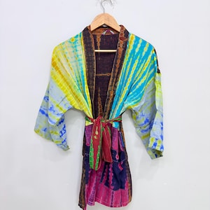 Puede incluir: Una bata kimono teñida con tintes en tonos amarillos, azules y rosas, colgada de una percha de madera. La bata presenta un estampado tie-dye con un cinturón rojo, verde y morado. La bata está hecha de una tela ligera.