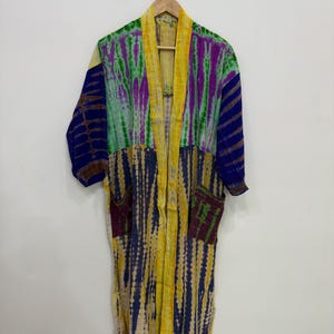 May include: A colorful tie-dye robe with a mix of vertical stripes in yellow, blue, green, and purple. The robe has two pockets and a yellow collar. The garment is hanging on a wooden hanger against a white wall.