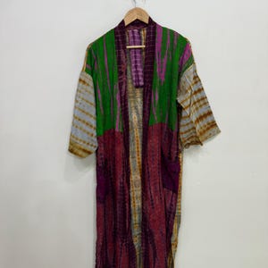 May include: A long, open-front robe with a tie-dye pattern in shades of green, purple, and brown. The robe is hanging on a wooden hanger against a white wall. The sleeves are a lighter color than the body of the robe.