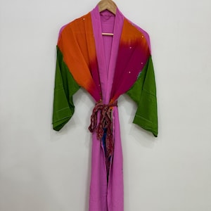 May include: A colorful robe with a tie belt, hanging on a wooden hanger. The robe features a vibrant mix of colors, including green sleeves, an orange and pink upper section, and a long pink body. The tie belt has a patterned design.