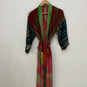 Könnte beinhalten: Ein langer, farbiger Robe in Rot-, Grün- und Dunkeltürkistönen, der an einem Holzbügel hängt. Die Robe hat einen Bindegürtel und ein gemustertes Design. Die Ärmel der Robe haben ein dunkelgrünes und goldenes Muster. Die Robe hängt vor einer weißen Wand.