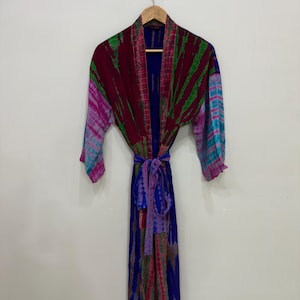 May include: A long, tie-dye robe in shades of blue, purple, green, and red hangs on a wooden hanger. The robe features long sleeves and a matching tie belt. The fabric has a flowing, lightweight appearance.