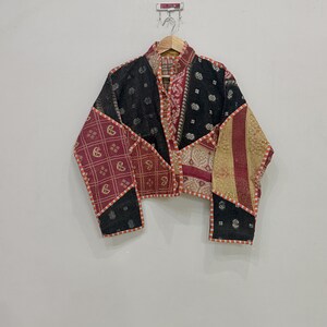 Può includere: Una giacca corta caratterizzata da un design patchwork con vari pannelli di tessuto in nero, rosso e oro. Il capo presenta motivi complessi tra cui paisley e forme geometriche, rifiniti con un bordo a righe arancione e bianco a contrasto.