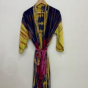 May include: A tie-dye robe in shades of yellow, blue, and pink hangs on a wooden hanger. The robe features a striped pattern and a matching belt. The garment is made of a lightweight fabric.
