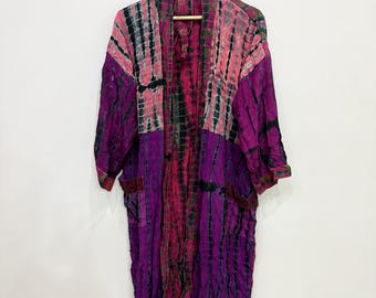 Puede incluir: Una bata estilo kimono de manga larga con un patrón tie-dye en tonos morado intenso, rosa brillante y negro. La prenda tiene la parte delantera abierta, un cuello ancho y dos bolsillos frontales, colgada de una percha de madera en una pared.