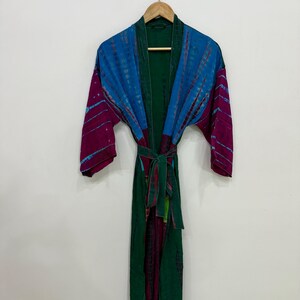 Può includere: Una vestaglia a maniche lunghe con un colore di base verde scuro e pannelli a contrasto in blu brillante e magenta. Il capo include una cintura in tessuto abbinata in vita e un motivo tie-dye sulle sezioni di colore blu e magenta.