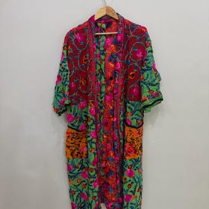 May include: A vibrant, embroidered kimono-style robe. The robe features a mix of red, green, orange, and pink floral patterns. It has short sleeves and two pockets. The robe is hanging on a wooden hanger.