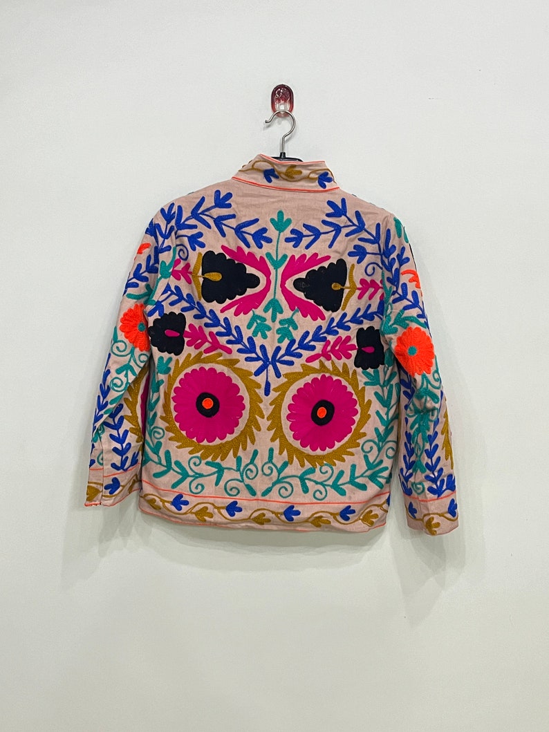 May include: A beige jacket with long sleeves and a high neck. The jacket is embroidered with colorful floral patterns in shades of pink, blue, green, orange, and black.