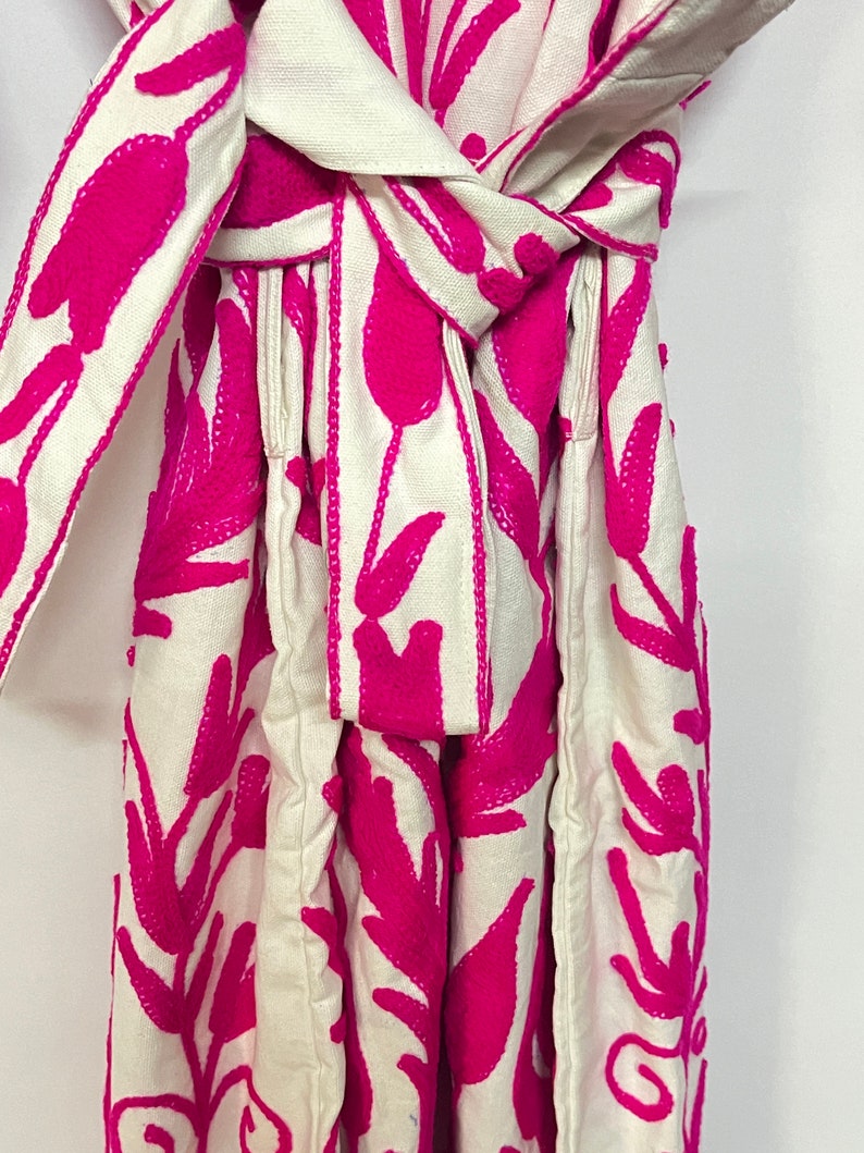 Puede incluir: Un vestido blanco con un patr&oacute;n de bordado floral rosa. El vestido tiene una faja que se ata a la cintura.