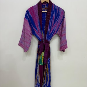 Puede incluir: Una bata teñida con tintes de azul, morado y rosa cuelga de una percha de madera. La bata presenta un diseño envolvente con un cinturón a juego. La tela tiene un aspecto texturizado y las mangas son de tres cuartos.