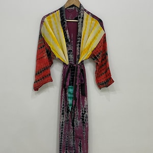 May include: A long, tie-dye robe with a vibrant color scheme. The robe features a mix of red, yellow, and purple hues, with black accents. It hangs on a wooden hanger against a plain white background. The robe has long sleeves and a matching belt.