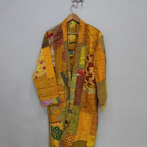 May include: A patchwork robe in shades of yellow, gold, and brown hangs on a wooden hanger. The robe is made of various fabric patches, stitched together with visible seams. The design includes floral and geometric patterns.
