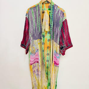 May include: A long-sleeved kimono robe featuring a vibrant patchwork of tie-dye patterns in shades of yellow, purple, pink, green, and orange. The garment includes an open front, a matching tie belt, and two front pockets with a floral print detail.