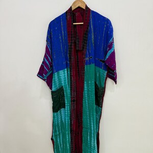 May include: A long-sleeved kimono robe featuring a tie-dye pattern in vibrant shades of blue, teal, and deep magenta. The garment includes a dark central panel, two front patch pockets, and an open front design, all hanging from a wooden hanger.