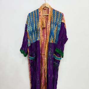 Può includere: Una vestaglia kimono a maniche lunghe con un vivace motivo tie-dye nei toni del viola, blu e arancione. Il capo presenta un'apertura frontale, un colletto ampio e due tasche anteriori con bordo verde. Il tessuto mostra un mix di righe verticali.