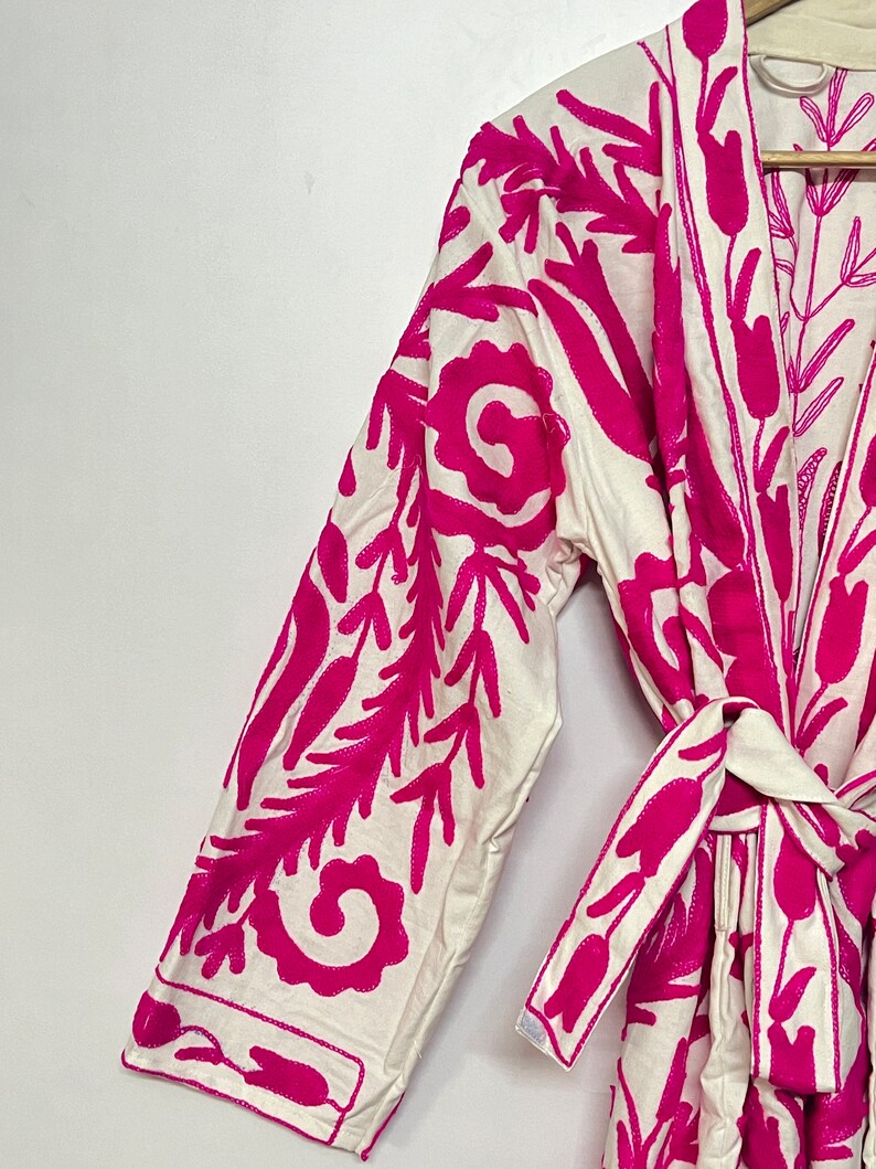 Puede incluir: Batista blanca con un dise&ntilde;o de bordado floral rosa. La bata tiene un cintur&oacute;n de lazo y mangas largas.