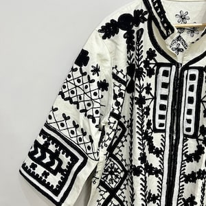 May include: White cotton shirt with black embroidered geometric patterns and floral designs. The shirt has a collar and short sleeves.