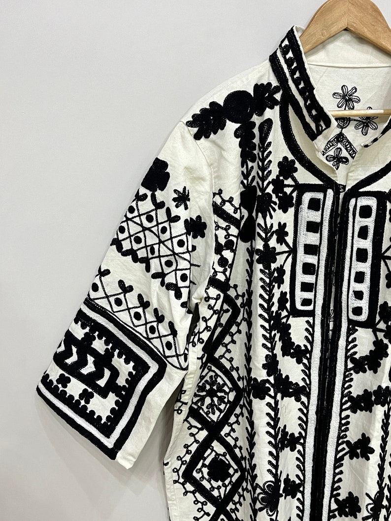 May include: White cotton shirt with black embroidered geometric patterns and floral designs. The shirt has a collar and short sleeves.
