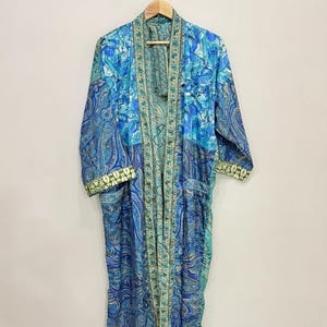 May include: A long-sleeved robe featuring a patchwork design in various shades of blue, teal, and green. The garment displays a mix of paisley and floral patterns, with a contrasting patterned trim along the front opening and sleeve cuffs.