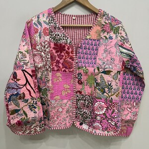 Op de afbeelding: Een roze gewatteerd jack met een patchwork-ontwerp met bloemenpatronen. Het jack heeft een V-hals en een knoopsluiting.