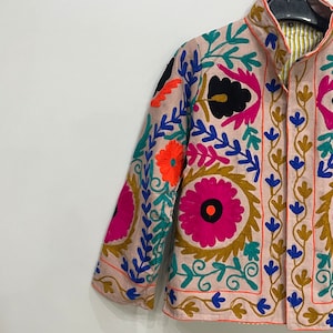 May include: A pink embroidered jacket with colorful floral patterns. The jacket has a mandarin collar and a button closure. The embroidery is detailed and intricate, featuring a variety of colors, including pink, blue, green, orange, and black.