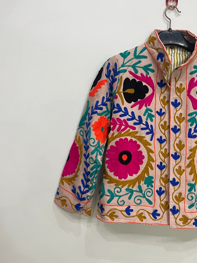 May include: A pink embroidered jacket with colorful floral patterns. The jacket has a mandarin collar and a button closure. The embroidery is detailed and intricate, featuring a variety of colors, including pink, blue, green, orange, and black.