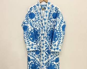 May include: A white robe with intricate blue floral embroidery. The long-sleeved robe is hanging on a wooden hanger. The embroidery features a repeating pattern of flowers and leaves.