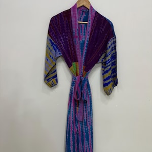 May include: A tie-dye robe in shades of purple, blue, and pink hangs on a wooden hanger. The robe features long sleeves and a matching tie belt. The fabric has a vibrant, colorful pattern.