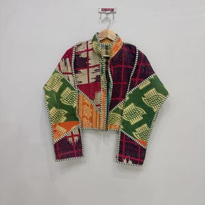 Könnte beinhalten: Eine langärmelige Jacke aus Patchwork-Stoff mit einer Vielzahl von Mustern und Farben, darunter Grün, Orange, Dunkelviolett und Creme. Das Kleidungsstück verfügt über einen Stehkragen, eine Vorderöffnung und sichtbare weiße Nähte.