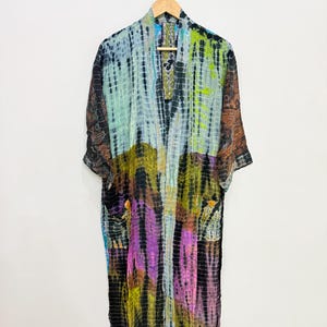 May include: A long-sleeved kimono robe featuring a multi-colored tie-dye pattern with shades of black, lime green, purple, and blue. The garment has an open front, a shawl collar, and two front pockets, hanging on a wooden hanger against a wall.