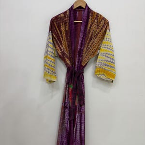 Puede incluir: Una bata de manga larga con un patrón de teñido anudado en tonos morado oscuro, marrón y amarillo. La prenda incluye un cinturón de tela a juego en la cintura, un borde morado sólido a lo largo de la abertura frontal y mangas anchas.