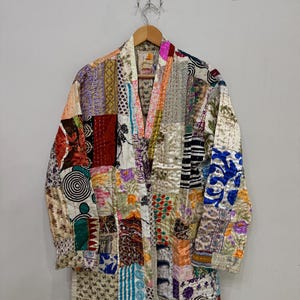 May include: A patchwork robe with a variety of colorful fabric squares. The robe is hanging on a wooden hanger. The design includes floral, geometric, and abstract patterns. The robe has long sleeves and a V-neck.