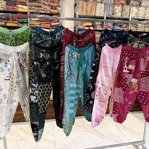 Könnte beinhalten: Eine Präsentation von bunten Patchwork-Hosen, die an einem Metallgestell hängen. Die Hosen weisen verschiedene Muster und Farben auf, darunter Weiß, Grün, Blau, Rosa und Burgunderrot. Die Hosen sind aus leichtem Stoff gefertigt und haben einen Kordelzug in der Taille. Im Hintergrund sind Regale mit Textilien zu sehen.