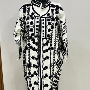 May include: A white long-sleeved jacket with black embroidery featuring floral and geometric patterns. The jacket has a mandarin collar and a front zipper closure.