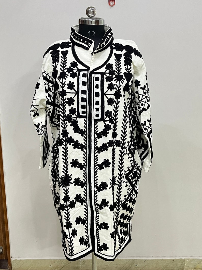 May include: A white long-sleeved jacket with black embroidery featuring floral and geometric patterns. The jacket has a mandarin collar and a front zipper closure.