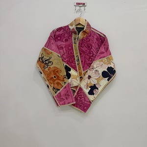 May include: A quilted jacket featuring a patchwork design of magenta, tan, and cream fabric panels with floral patterns. The garment has a high collar, long sleeves, and visible white stitching along the seams and edges, hanging on a wooden hanger.